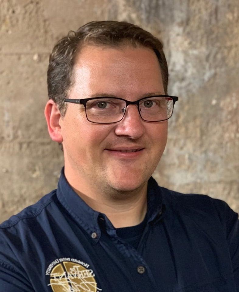 A smiling man with glasses wearing a dark blue Howard Payne University shirt against a textured background. | HPU